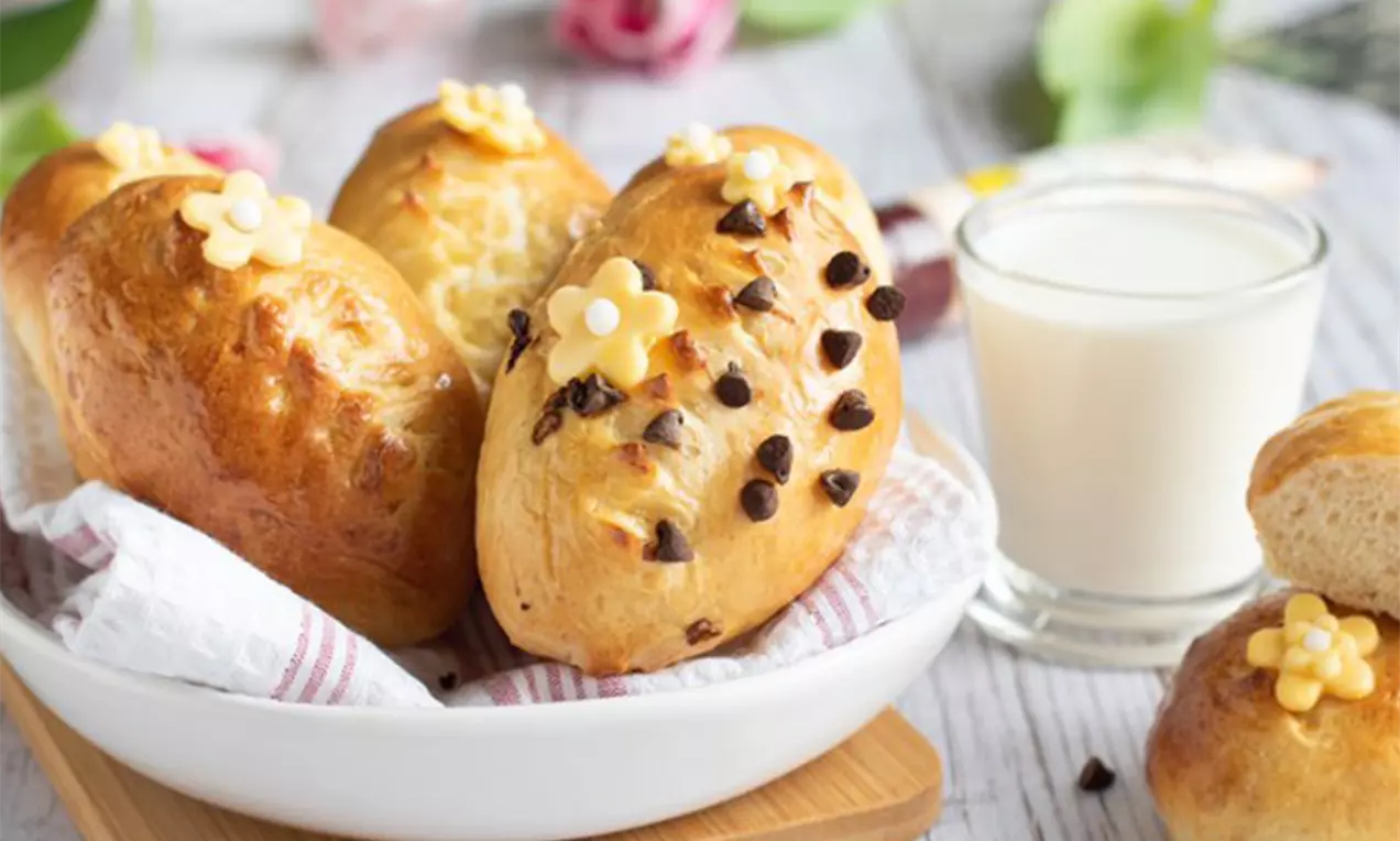 Goldbraune, glänzende Milchbrötchen mit Schokotropfen und gelben Zuckerdekor-Blumen, angerichtet in einer weißen Schale auf Holzbrett
